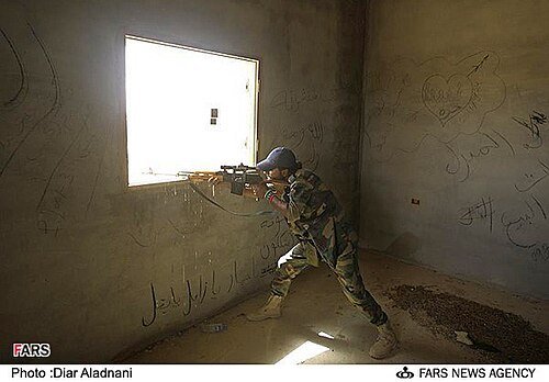 A man in camouflage trousers and a cap aims a rifle out of a square window in a bare concrete room with writing on the walls