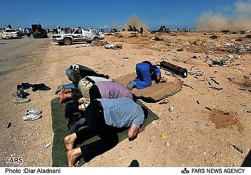Several men praying on mats on stony ground beside a road, with pickup trucks and rising smoke in the background