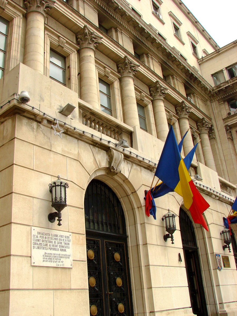 A stone-faced corner of the former Central Committee building in Bucharest with a balcony above, Romanian flags and a plaque on the wall