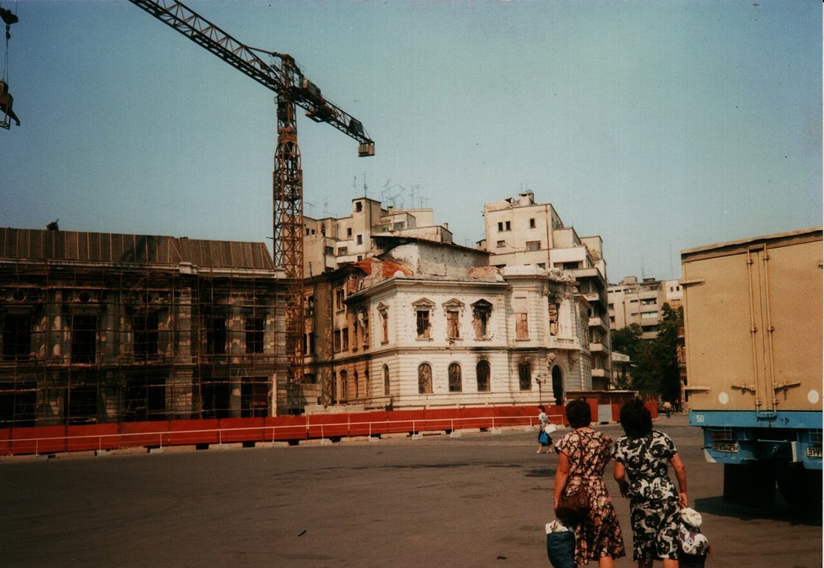 Damaged buildings behind a red hoarding and scaffolding, with a tower crane and two women walking across an open square