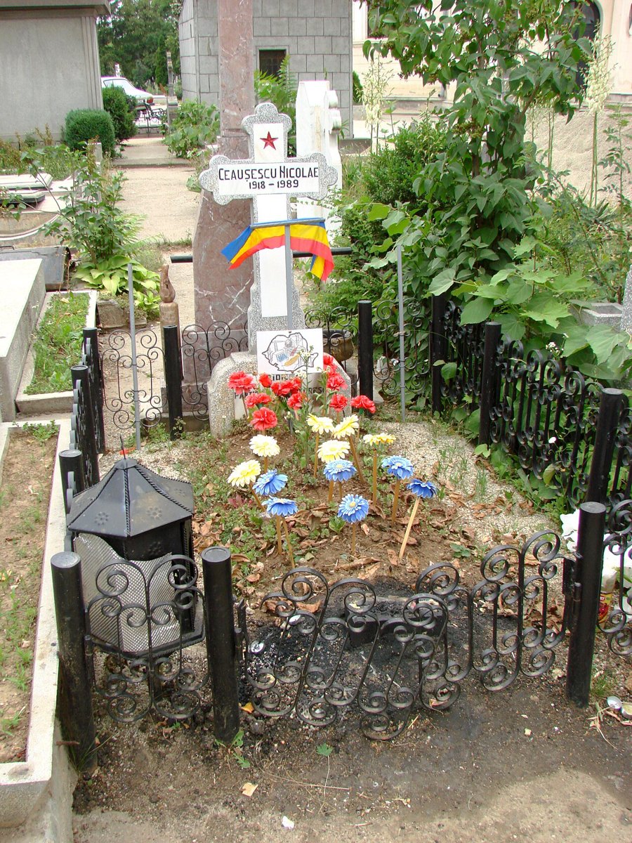 A small fenced grave in a cemetery with a white cross reading Ceausescu Nicolae 1918-1989, a red star, a Romanian flag and artificial flowers