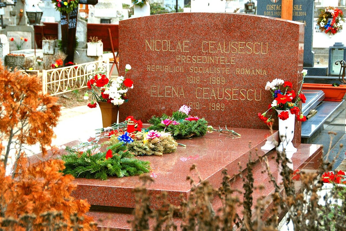 A red granite gravestone with gold lettering naming Nicolae Ceausescu, President of the Socialist Republic of Romania, 1918-1989, and Elena Ceausescu, 1919-1989, with flowers and wreaths on it