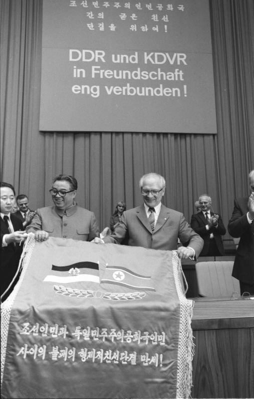 Kim Il Sung and Erich Honecker holding a banner with the East German and North Korean flags, under a sign in German and Korean reading 'DDR und KDVR in Freundschaft eng verbunden'