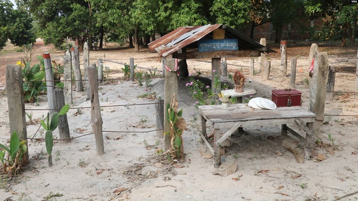 A fenced sandy plot with a small tin-roofed shelter, a low wooden table holding offerings and a red box, and a blue sign in Khmer script