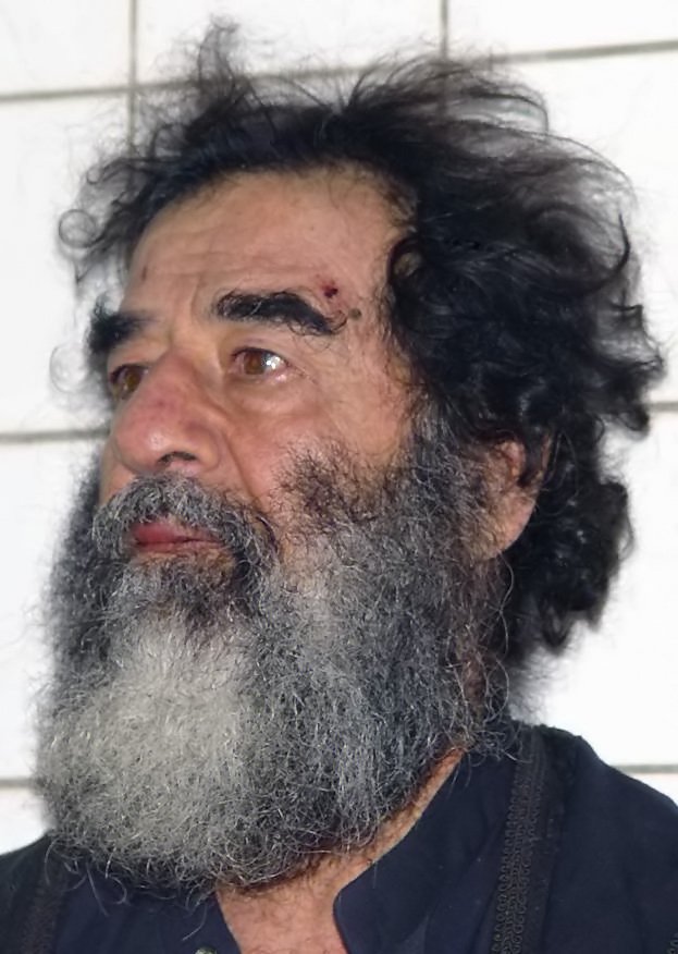 Close-up of Saddam Hussein with long unkempt hair and a grey beard, with a small mark above the left eyebrow, against a white tiled wall