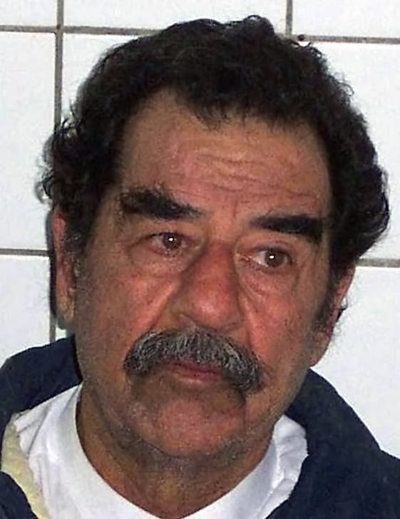 Close-up of Saddam Hussein clean-shaven apart from a grey moustache, with short dark hair, in a blue jacket against a white tiled wall
