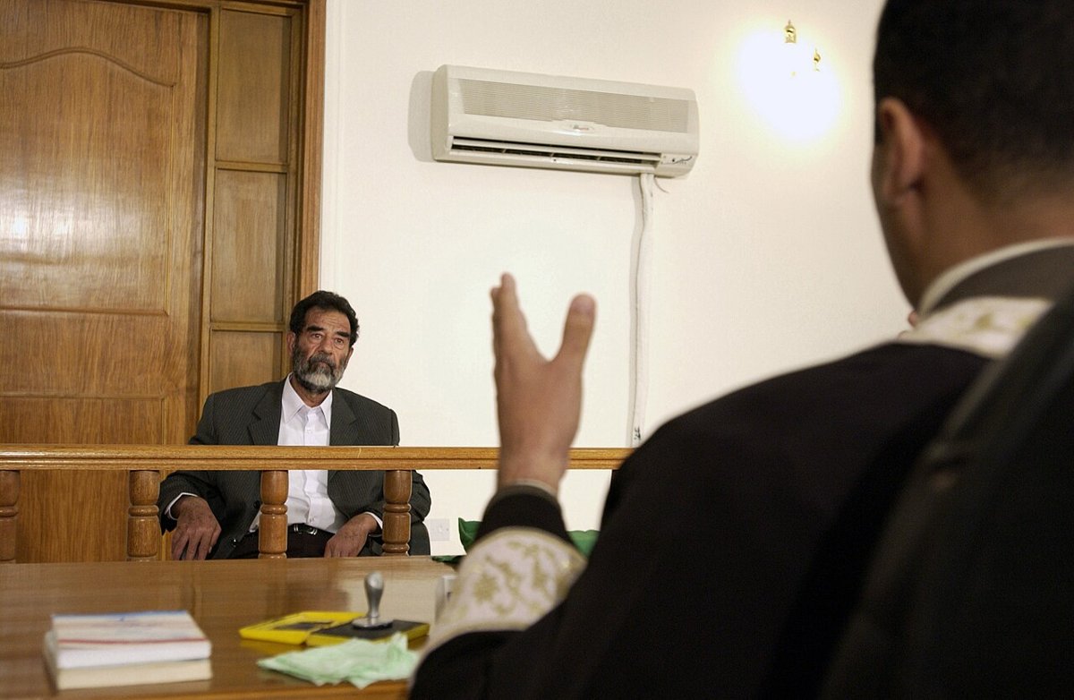 Saddam Hussein in a dark suit and white shirt seated behind a wooden rail, facing a judge in a robe who is seen from behind with one hand raised