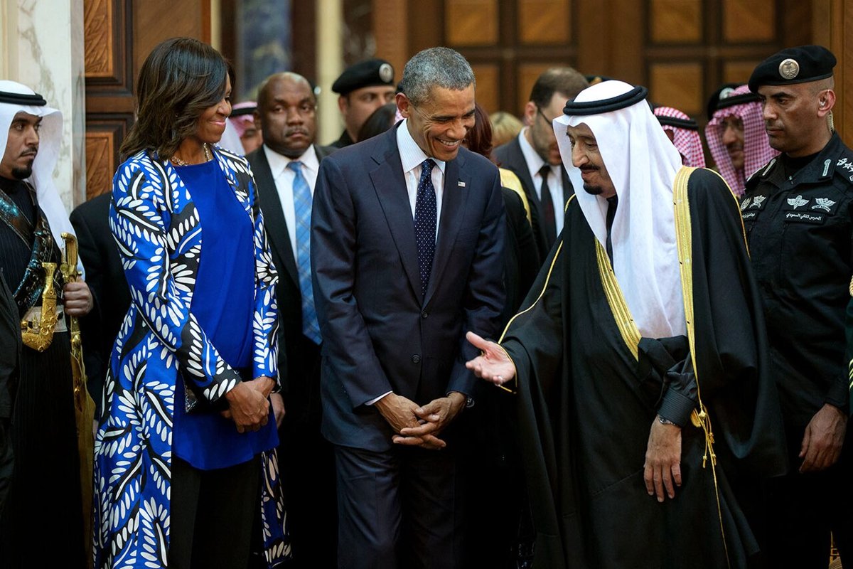 An older man in a black cloak and white headdress gesturing towards a smiling man in a dark suit, with a woman in a blue patterned coat and uniformed guards beside them in a panelled hall