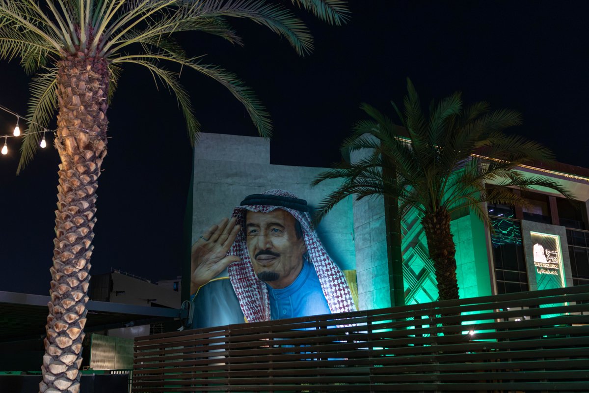 A large illuminated image of an older man in a red-and-white headdress, raising one hand in salute, on the wall of a building at night between two palm trees