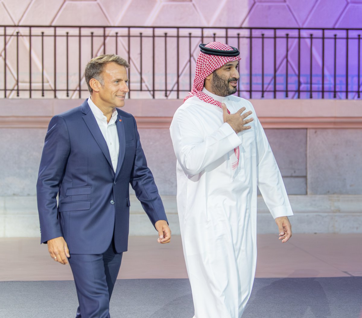 Two men walking side by side outdoors in evening light, one in a dark suit and open-necked shirt, the other in a white thobe and red-and-white headdress with a hand on his chest