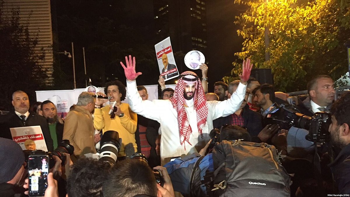 At night, a man in a white robe and red-and-white headdress raises both hands, painted red, among photographers and demonstrators holding placards with Jamal Khashoggi's photograph and Arabic text