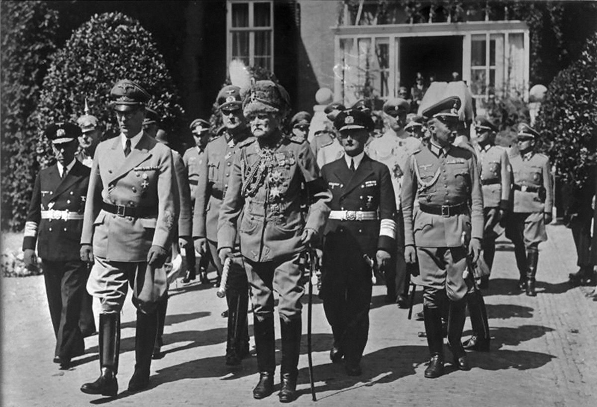 Uniformed German officers and officials walking in a column along a paved path beside clipped hedges; the man at the centre wears an old Hussar uniform with a fur busby and a black armband