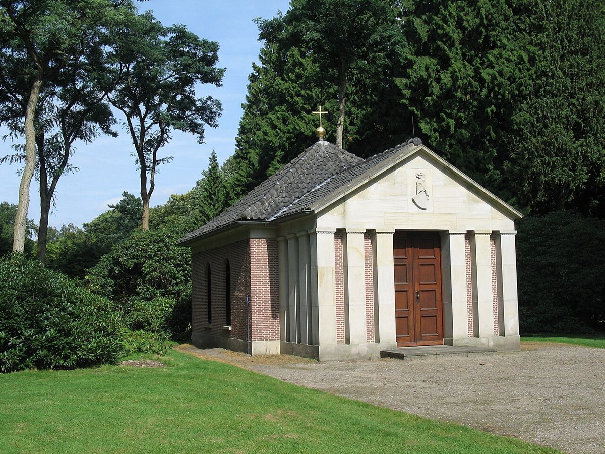 A small classical temple of pale stone and red brick with a pedimented porch, a pair of wooden doors and a gold orb and cross on its roof, among tall trees