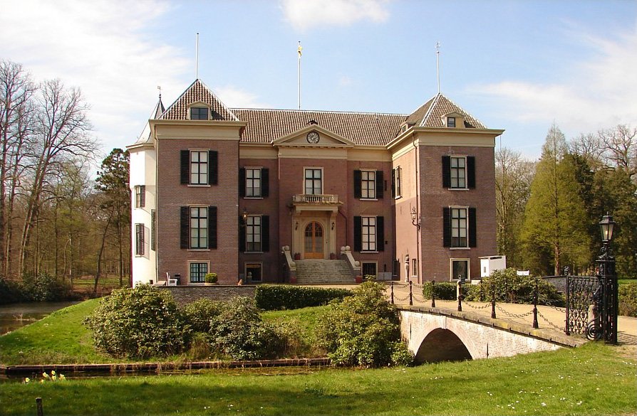 A two-storey brick manor house with shuttered windows, stepped gables and a front stair, seen across a moat and a small stone bridge