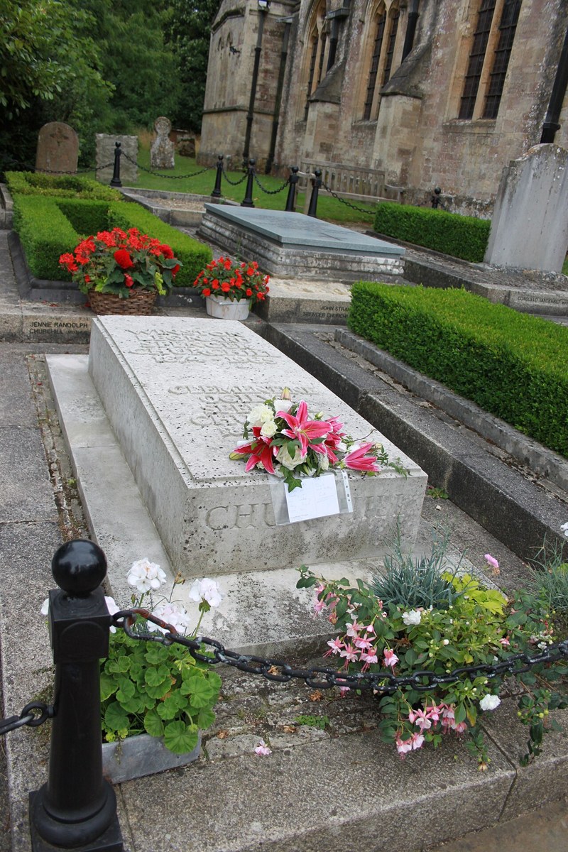 A low pale stone tomb with flowers on it, marked CHURCHILL, in a churchyard with box hedges beside a stone church