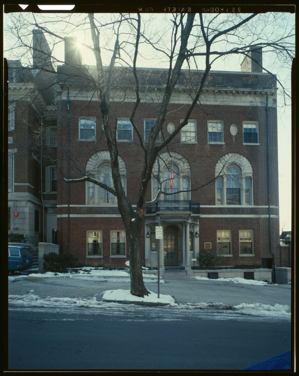 The brick front of a three-storey Georgian-style town house with arched upper windows and a small balcony over the door, seen through a bare tree in winter