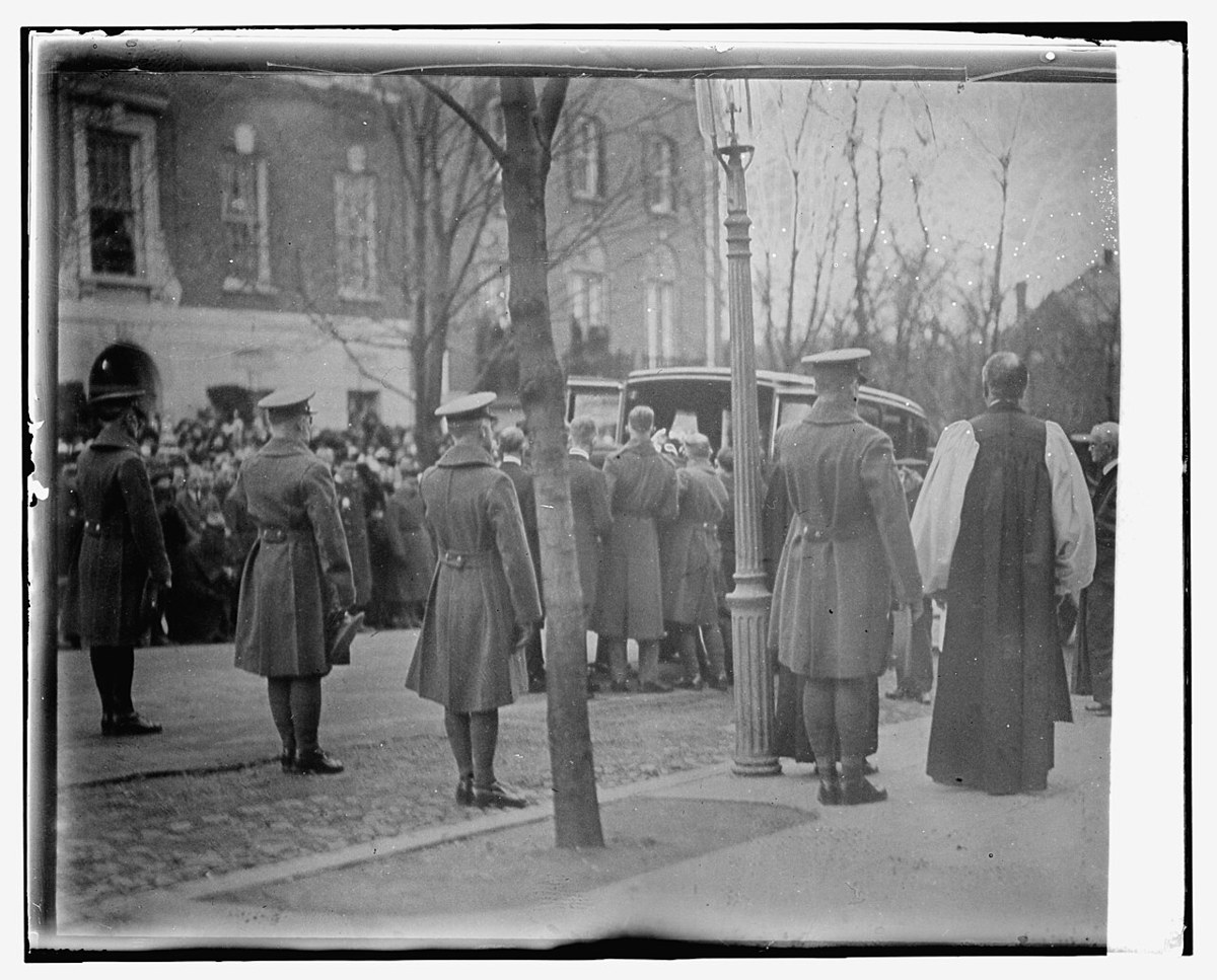 Soldiers in greatcoats and a clergyman in white surplice stand in a Washington street as people in overcoats gather around a hearse in the background