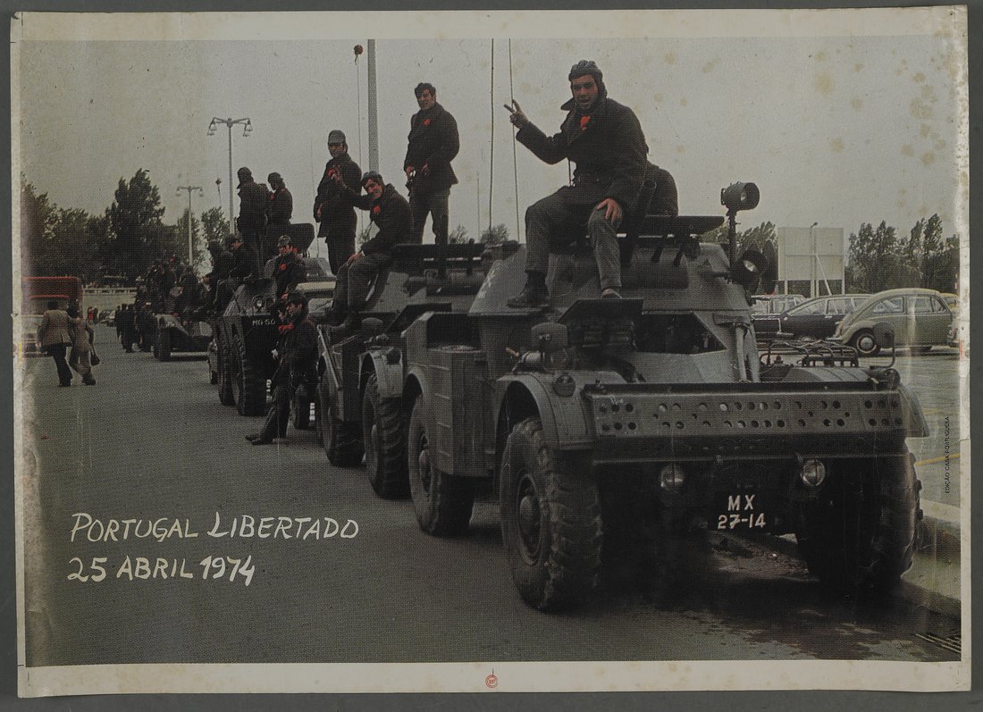 Armoured cars in a line on a road with soldiers standing on them, red carnations on their uniforms, one raising a hand in a V sign, with the hand-lettered words Portugal Libertado 25 Abril 1974