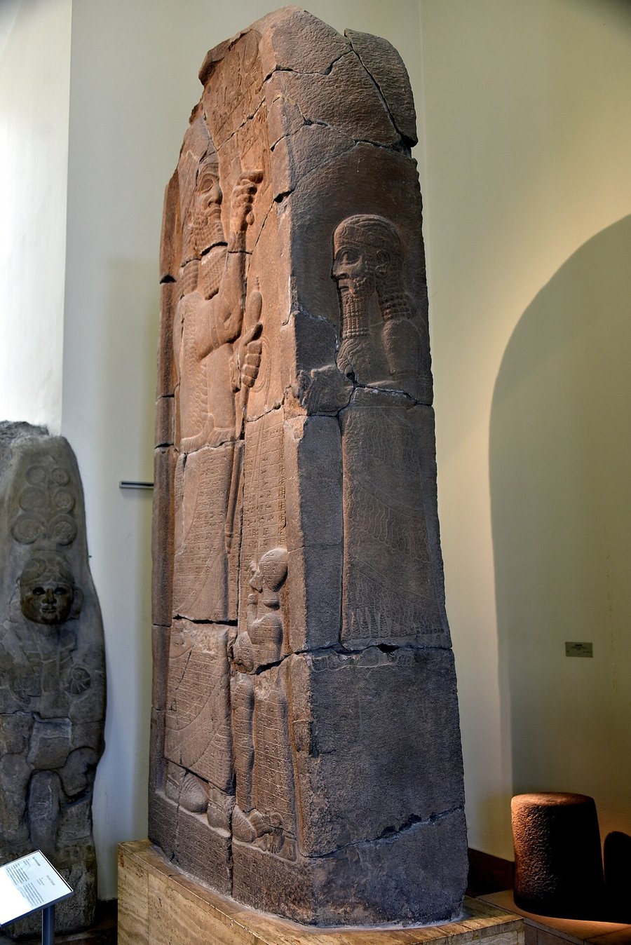 A tall carved stone stele in a museum hall, with a large standing king in relief and two small figures at his feet