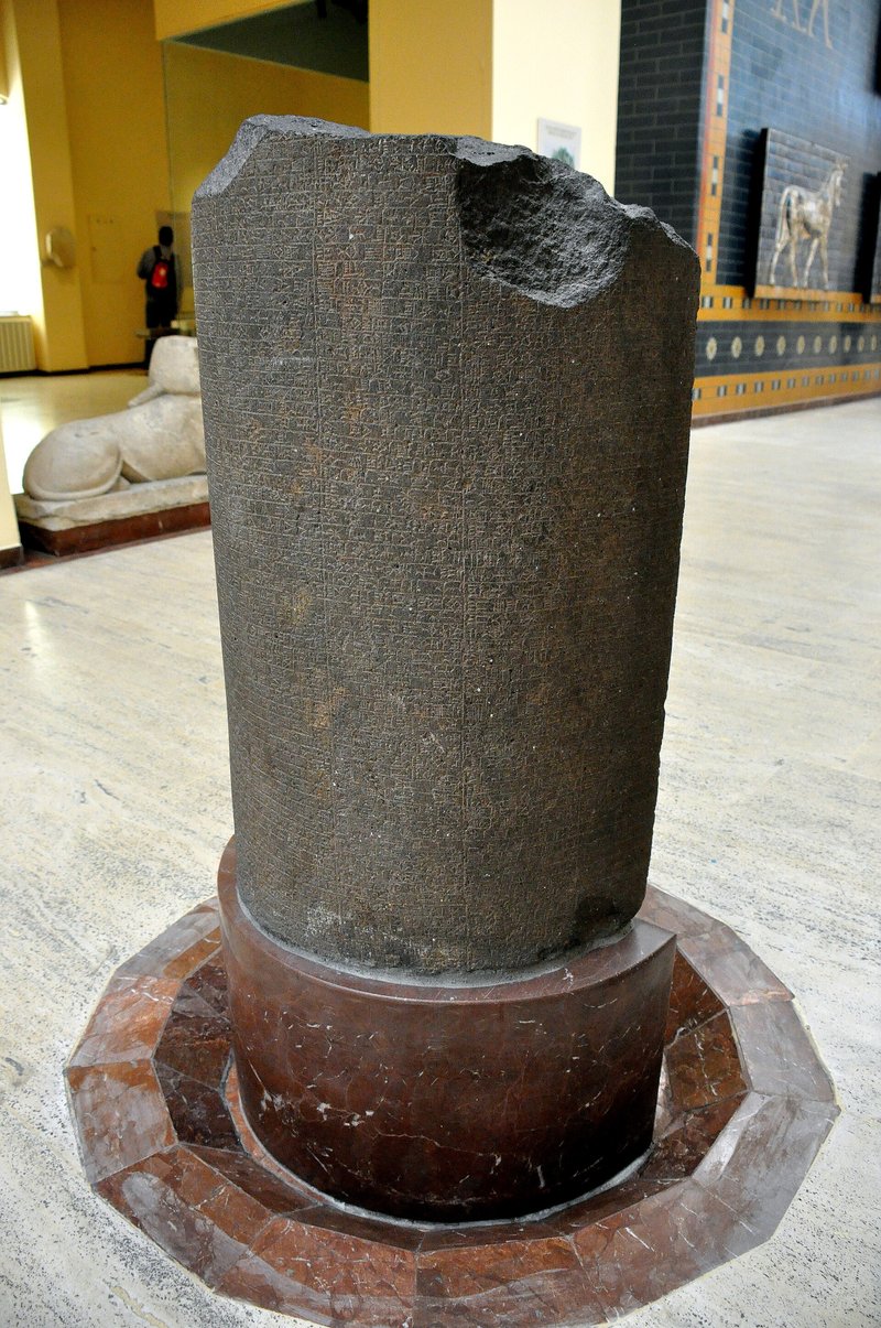 A dark granite stele standing on a round stone base in a museum hall, its broken top at the right and its face covered in lines of cuneiform
