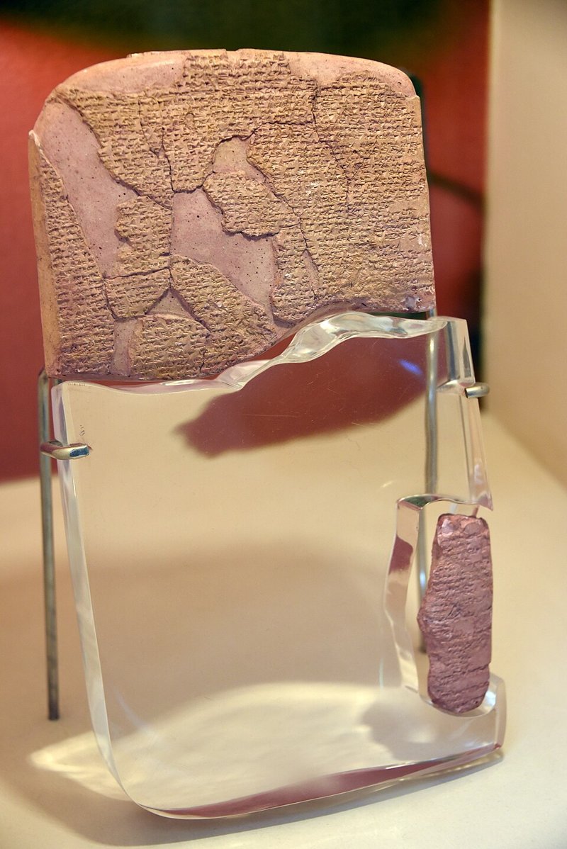 A cracked reddish clay tablet covered in cuneiform wedges, in two fragments, set on a clear plastic stand.