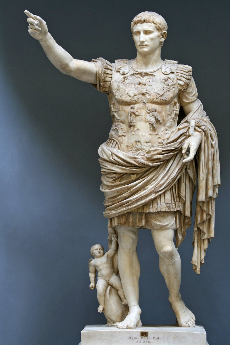 Marble statue of Augustus in a muscled cuirass, right arm raised, a small figure at his right leg, in a museum
