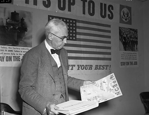 Elmer Davis at a press conference on 6 March 1943, examining Axis propaganda publications in front of OWI posters