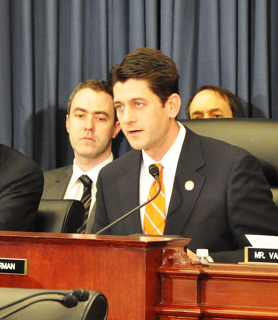Representative Paul Ryan speaking into a microphone at a House committee table, with two men seated behind him