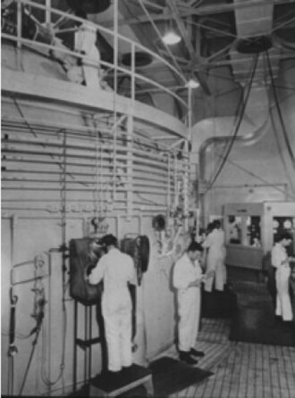 Black-and-white photograph of workers in white coats at glove ports beside the curved wall of a very large sphere inside a building