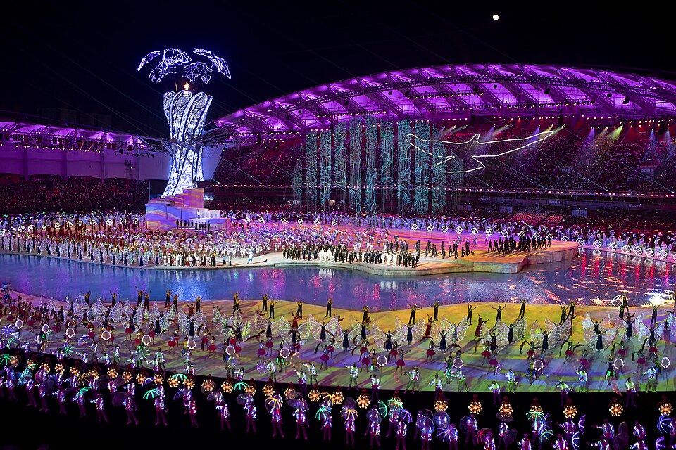 Night view of the opening ceremony of the 2019 Military World Games in Wuhan: a lit stadium roof, a tall illuminated cauldron and performers in illuminated costumes on a stage