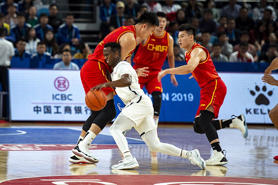 Two players in red China jerseys defend against a player in white as he dribbles a basketball, in an arena with advertising boards