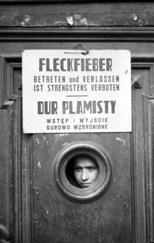 A printed German and Polish typhus notice on a wooden door in the Warsaw Ghetto, with a person looking out through a round window in the door below it